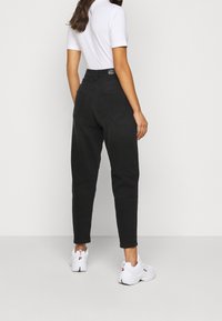 Kvinna som står med ryggen vänd, iklädd högmidjade svarta jeans, en vit kortärmad topp och vita FILA sneakers mot en enfärgad bakgrund.