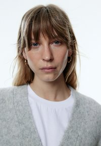 Grijze gebreide cardigan over een witte T-shirt, met een diepe V-hals. De textuur lijkt zacht; het model heeft steil haar en kleine hooporen.