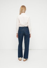 Vaqueros de mezclilla azul con cintura alta, corte de pierna ancha y dos bolsillos traseros. Combinados con una camisa blanca de manga larga con botones.