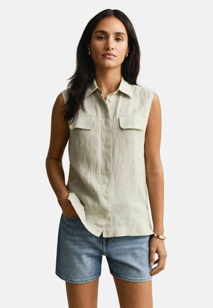 Frau mit langen dunklen Haaren, die ein ärmelloses beige Hemd mit Knöpfen und blaue Jeansshorts trägt, mit einer Hand in der Tasche.