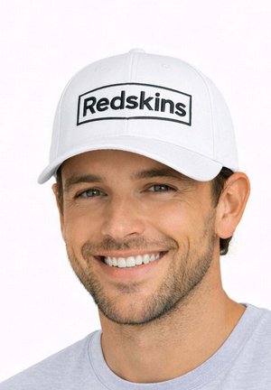 Homme souriant aux cheveux courts châtains et à la barbe, portant une casquette blanche avec l’inscription « Redskins » et une chemise gris clair sur un fond uni.