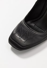 Černý, lesklý lodičkový podpatek s kulatou špičkou, na kterém jsou na nártu embosované loga "NY" a "Calvin Klein". Hladký povrch.