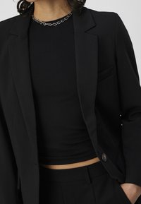 Blazer noir ajusté avec un seul bouton, porté sur un haut noir côtelé. Caractérisé par une coupe slim et une texture lisse. Design minimaliste.