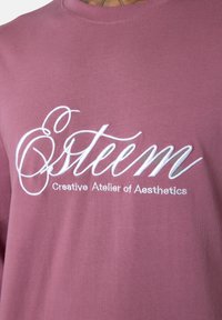 Staubiges rosafarbenes Gewebe mit weißem, gesticktem Text "Osteem" und kleinerem Text darunter mit der Aufschrift "Kreatives Atelier der Ästhetik" auf Kleidung.