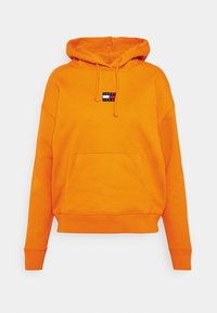 Sudadera con capucha naranja con bolsillo delantero, capucha ajustable con cordón y un pequeño parche negro con la marca. Material de algodón suave, hombros caídos.