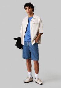 Lichtbeige overhemd met korte mouwen over een blauw T-shirt, gecombineerd met blauwe denim short en crèmekleurige sneakers met zwarte accenten. Witte sokken zichtbaar.