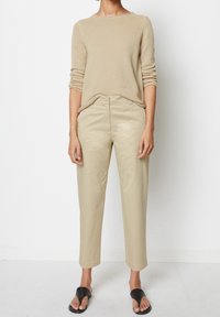 Pull beige en maille à manches longues et col montant. Pantalon fuselé kaki clair avec poches latérales. Combiné avec des sandales noires à enfiler.