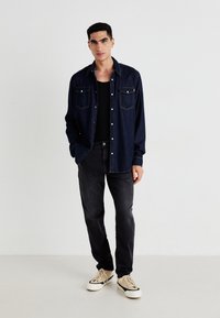 Laivastonsininen farkk Shirt, jossa on kaksi rintataskua, musta aluspaita, tummat farkut ja kermaiset tennarit. Paidassa on valkoiset napit ja näkyvät ompeleet.
