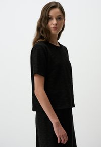 Schwarzes, kurz geschnittenes T-Shirt aus strukturiertem Stoff mit kurzen Ärmeln und einem runden Halsausschnitt, getragen mit einem passenden schwarzen Kleidungsstück.