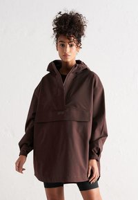Anorak marrón impermeable con capucha, mangas largas, bolsillo frontal y un logo reflectante. Textura suave, ajuste holgado y diseño minimalista.