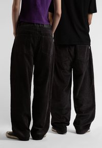 Pantaloni di velluto a coste neri larghi con tasche, caratterizzati da una texture a righe verticali. Indossati con una maglietta viola e nera, che mostra colori a contrasto.