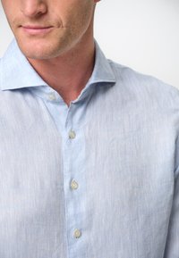 Camicia di lino azzurro chiaro con colletto classico, caratterizzata da un tessuto strutturato e tre bottoni sulla patta, parzialmente visibili.
