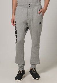 Grå sweatpants med tvåknappslinning, sidofickor och svart Nike-logotyp längs vänster ben. Tillverkade av en mjuk bomullsblandning.