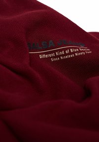 Tela burdeos con una textura suave que presenta el logotipo y el eslogan "SALSA JEANS" en una impresión contrastante, destacando la herencia del denim desde 1994.