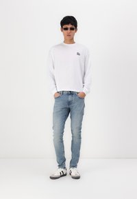 White long-sleeve shirt with black text, light blue jeans, and black and white sneakers with gray accents; model stands with hands in pockets.