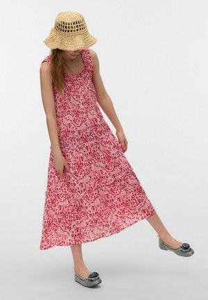 Pige iført en pink mønstret ærmeløs kjole, stråhat, der dækker hendes ansigt, og grå flade sko med sorte blomsterdetaljer, løfter det ene ben.