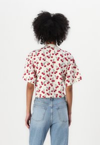 Cămașă scurtă, crop, în crem cu un model de cireșe roșii. Țesătură moale cu o croială lejeră și decolteu rotund, combinată cu blugi cu talie înaltă.