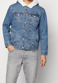 Blaues Denim-Jackett mit einer weichen, weißen Fleece-gefütterten Kapuze, knöpfbarem Verschluss, Brusttaschen und klassischem Schnitt. Getragen mit grauen Jeans.