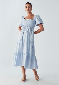 Svetlo modra maksi obleka z nabranimi rokavi, smukano bodico, večplastnim krilom in razširjenim robom. Narejena iz mehkega, lahkega materiala.