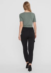Pantaloni cargo neri con vita elasticizzata e polsini alla caviglia, abbinati a una maglietta corta verde oliva. Il modello è visto da dietro.