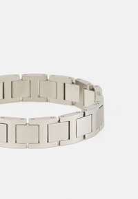 Bracelet en acier inoxydable argenté avec des maillons rectangulaires à finition brossée. Texture lisse avec un éclat subtil et un matériel de connexion visible.