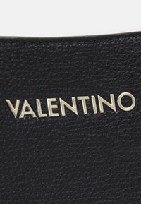 Svart teksturert lær med en metallisk gul "VALENTINO"-logo tydelig fremhevet, som viser en slank og moderne design.