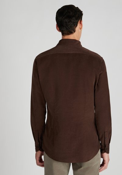 Man die met zijn rug naar de camera staat, gekleed in een lang mouw donkerbruine corduroy overhemd en een lichtbeige broek tegen een effen achtergrond.