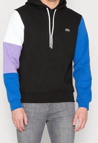 Felpa con cappuccio con un corpo nero, dettagli bianchi e viola sulle maniche e polsini blu. Presenta una tasca a marsupio frontale e un cappuccio con cordino.