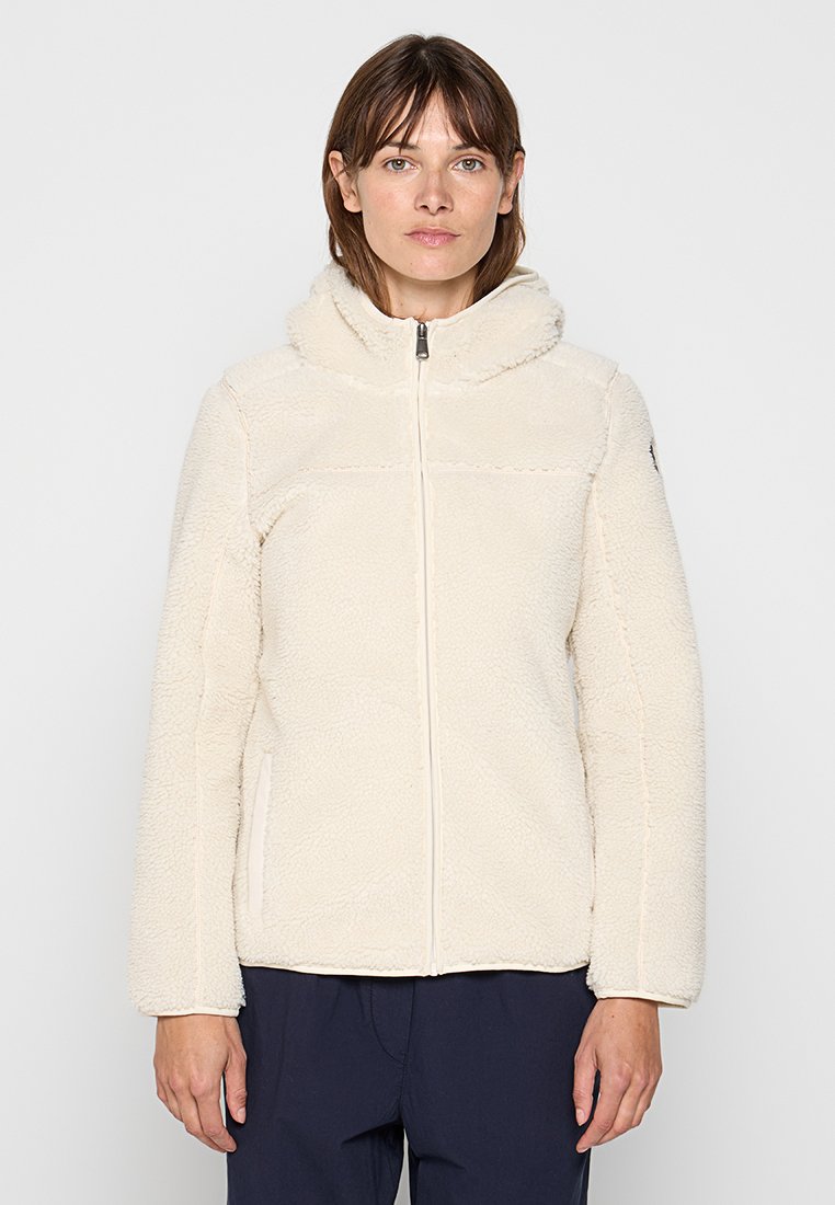 Femme portant une veste zippée en peluche couleur crème avec une capuche et un pantalon navy, se tenant devant un fond clair.