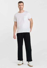 Man staat in een wit overhemd met korte mouwen en borstzak, zwarte broek met manchet en witte sneakers op een effen achtergrond.