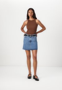 Bruine mouwloze top, blauwe denim minirok, zwarte riem met zilveren gesp en zwarte gepatineerde Mary Jane schoenen. Effen achtergrond.