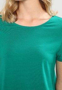 Vrouw in een groene crewneck T-shirt met korte mouwen en blond schouderlang haar.