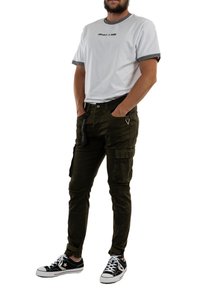 Camiseta blanca con ribete gris y logo, pantalones cargo verde oliva con bolsillos laterales, y zapatillas negras con detalles en blanco.