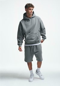 Grå hettegenser med lomme foran, kombinert med matchende shorts. Modellen har på seg hvite joggesko, som viser en avslappet athleisure-stil.