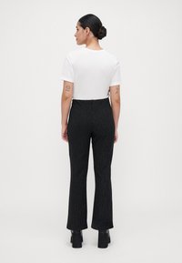 Femme avec les cheveux en chignon portant un top blanc à manches courtes, un pantalon noir à rayures et des chaussures à talons noires se tenant de dos contre un fond blanc.