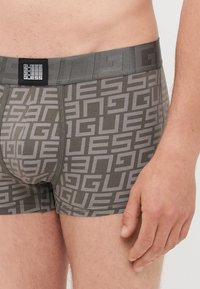 Sötét szürke boxeralsók, amelyek ismétlődő világosszürke "GUESS" logó mintával rendelkeznek. Gumis derékpánt, texturált felülettel.