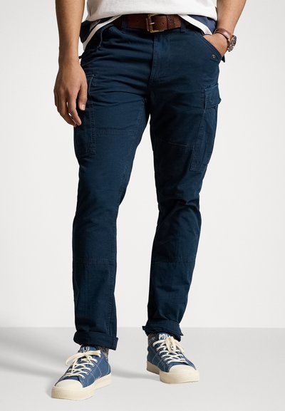 Pantaloni cargo navy cu numeroase buzunare laterale, realizați dintr-un material rezistent. Purtați cu o curea maro și adidași din pânză alba, cu accente albe.