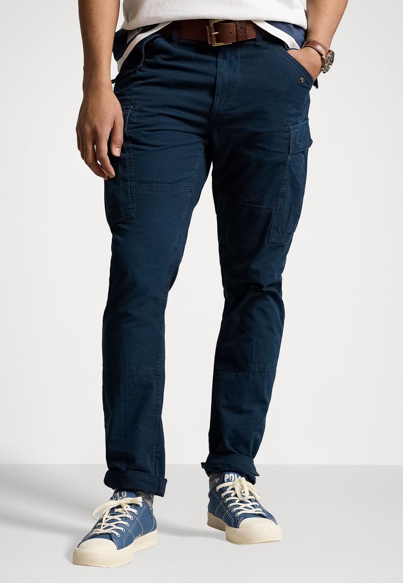 Navy cargo pants with multiple side pockets, made from sturdy fabric. Worn with a brown belt and blue canvas shoes, featuring white accents.