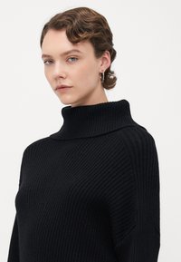 Ung person med kort, vågigt hår, som bär silverfärgade hoops örhängen och en svart ribbad polotröja, mot en enkel vit bakgrund.