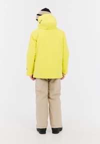 Heldere gele waterdichte jas met een capuchon, gecombineerd met lichtbeige broek en zwarte schoenen, gezien van achteren.