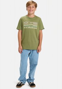 Zelena bombažna majica s prikazom gore in napisom "Quiksilver". Kombinirana s svetlo modrimi jeans hlačami in črnimi supergami.