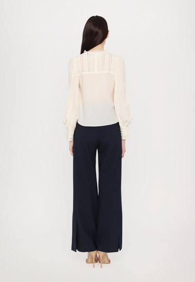 Ivory-blouse, jossa on puffihuput, pitsiyksityiskohtia selässä, yhdistetty tummansinisiin leveälahkeisiin housuihin, joissa on sivuhalkiot. Kirkas, sileä kangas.