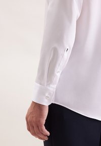 De hand van een man naast de manchetknoop van een witte blouse met lange mouwen, versierd met een knoop en een kleine zwarte rozenborduursel op de mouw, gecombineerd met donkere broek.