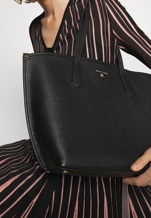 Osoba oblečená v černo-růžovém pruhovaném oblečení drží velkou černou texturovanou tašku Michael Kors s dvojitými uchy.