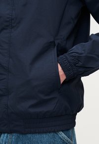 Άνδρας με σκούρο μπλε μπουφάν με ελαστικές μανσέτες, με το χέρι μερικώς στην πλαϊνή τσέπη, φορώντας μπλε τζιν παντελόνι.