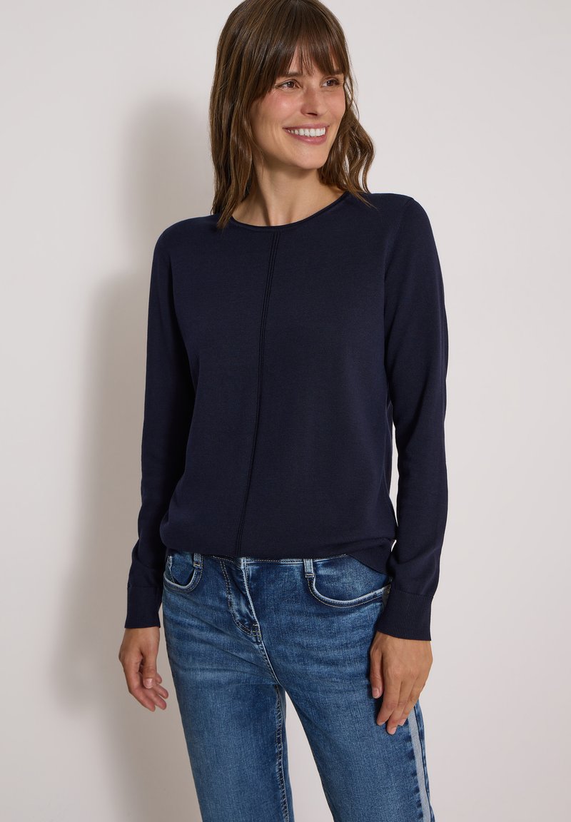 Femme souriante portant un pull bleu marine à manches longues avec un détail de couture centrale et un jean en denim bleu sur un fond uni.