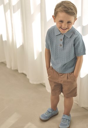 Polo en tricot bleu clair avec trois boutons marron, associé à un short kaki. Baskets bleues avec bandes Velcro. Debout sur un sol clair.