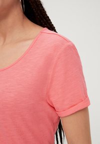 T-shirt léger de couleur corail avec un décolleté en forme de scoop, des manches courtes roulées et un tissu doux légèrement texturé.