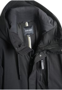 Chaqueta negra con capucha, que presenta una textura suave, bolsillos con cremallera y una etiqueta de marca en el interior, acentuada por una franja central.