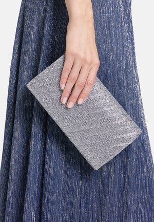 Sølvmetallic clutch med en tekstureret overflade holdt i en hånd, sat mod en mørkeblå plisseret stofbaggrund.